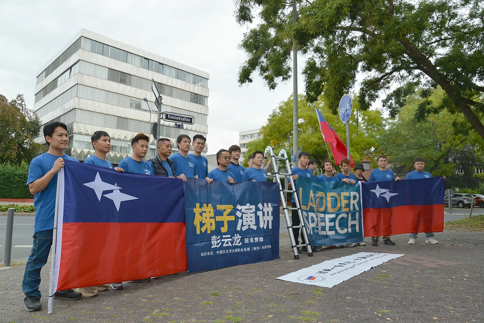 Members of the German branch in blue T-shirts hold a party flag and the “梯子演讲 LADDER SPEECH” banner at the street corner opposite the consulate.
