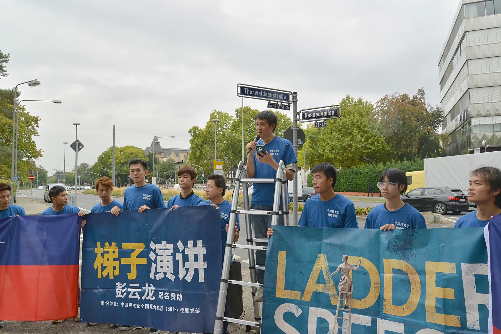 Members in blue T-shirts hold the banner and a party flag at the corner; a speaker stands on the ladder behind them.