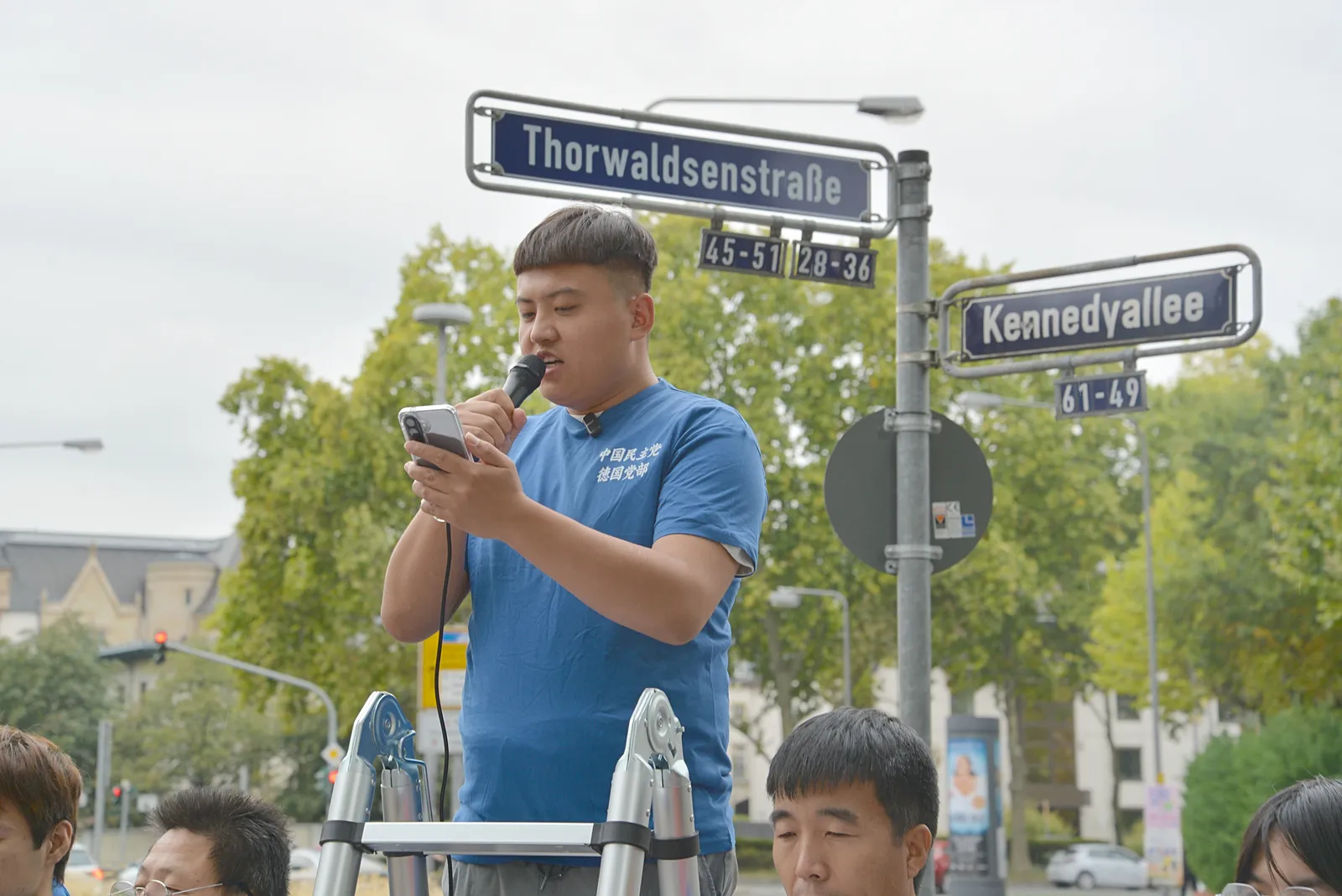A speaker in a blue T-shirt holds a microphone and a phone on the ladder, with the Thorwaldsenstraße and Kennedyallee street signs behind him.