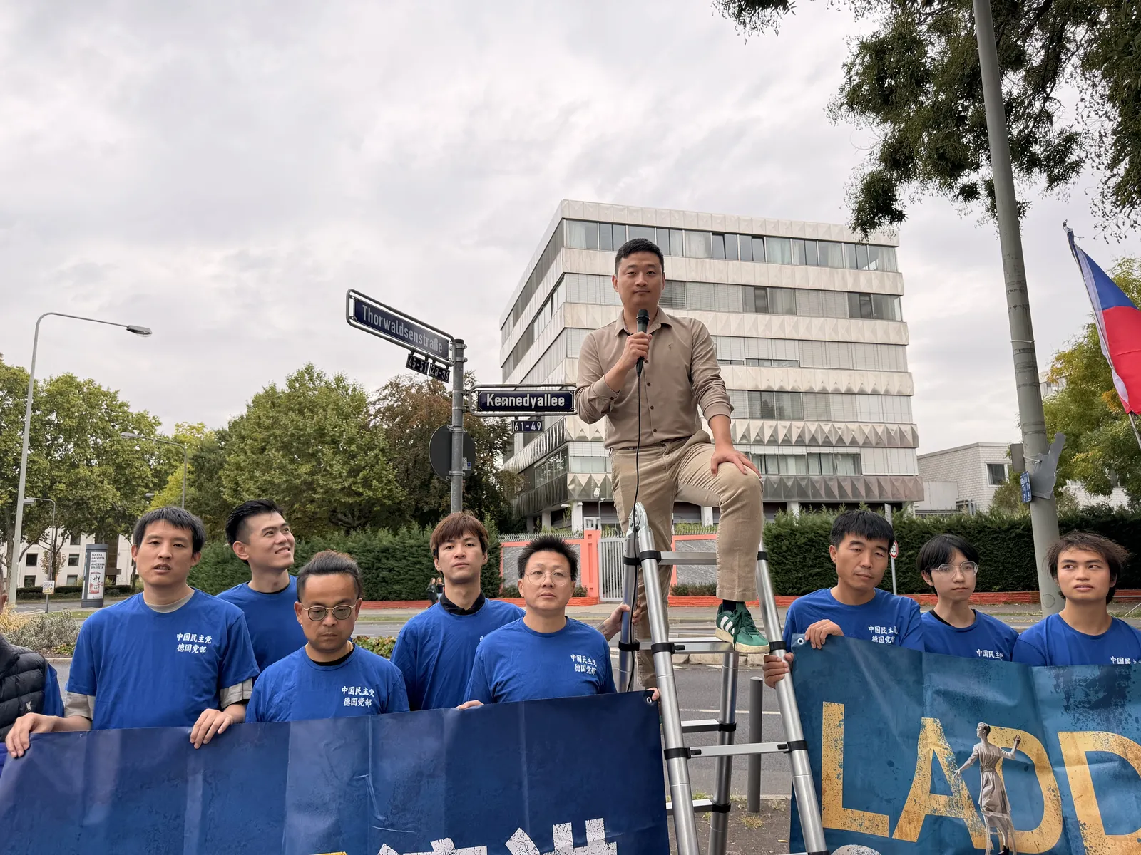 A speaker sits at the top of the ladder with a microphone while members in blue T-shirts stand below holding the banner.
