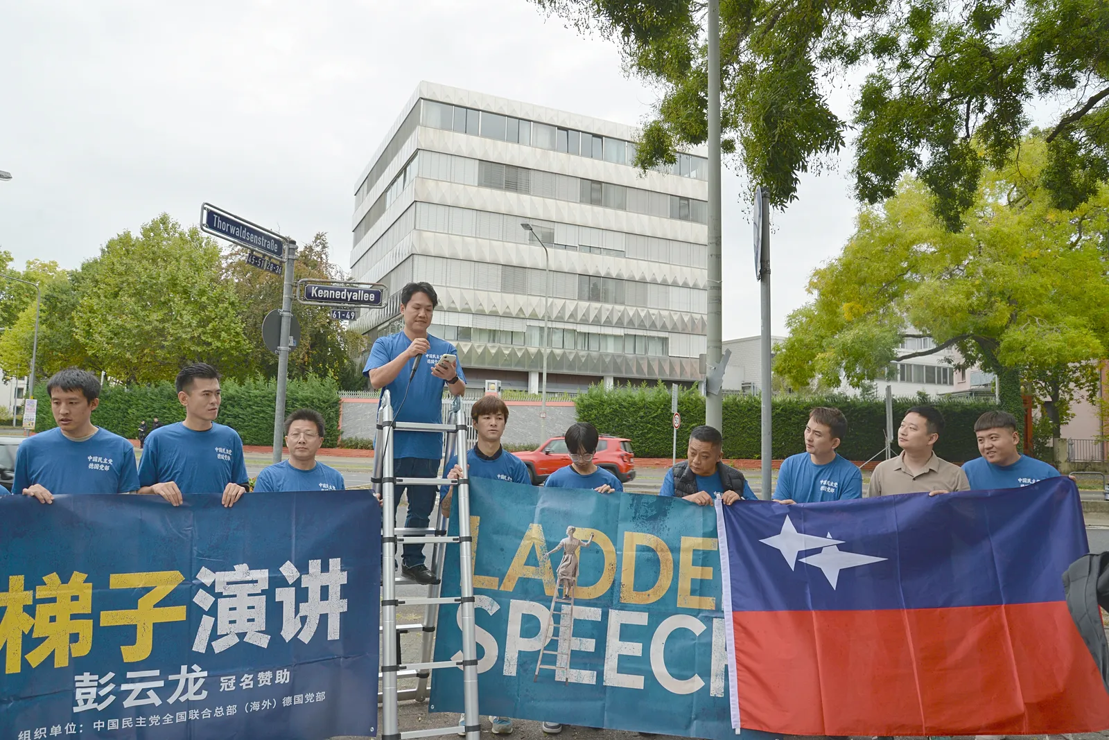 Members hold a party flag and the banner at the street corner, with a speaker on the ladder and the consulate building behind.