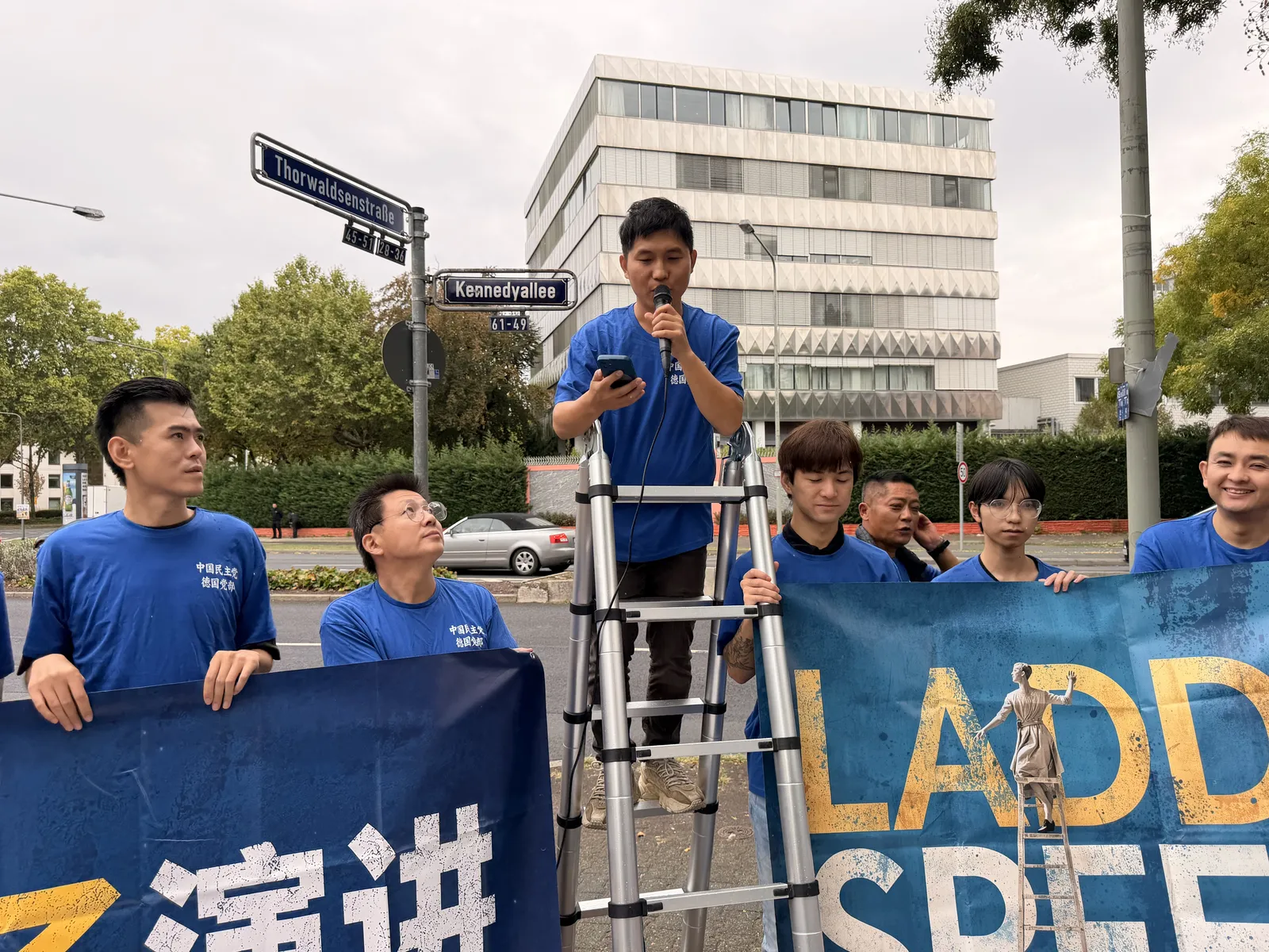 A speaker in a blue T-shirt speaks into a microphone on the ladder while members hold the banner and look up.