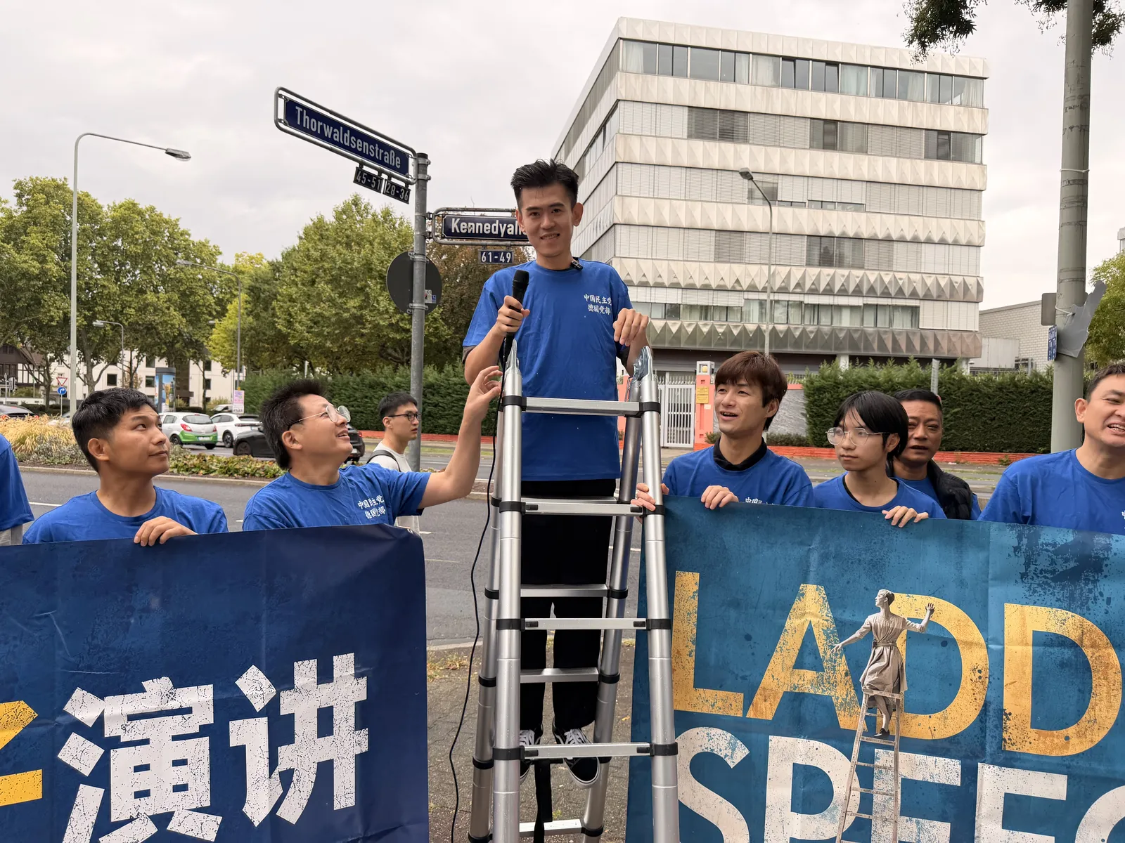 A speaker stands on the ladder with members in blue T-shirts around its foot and the banner beside them.