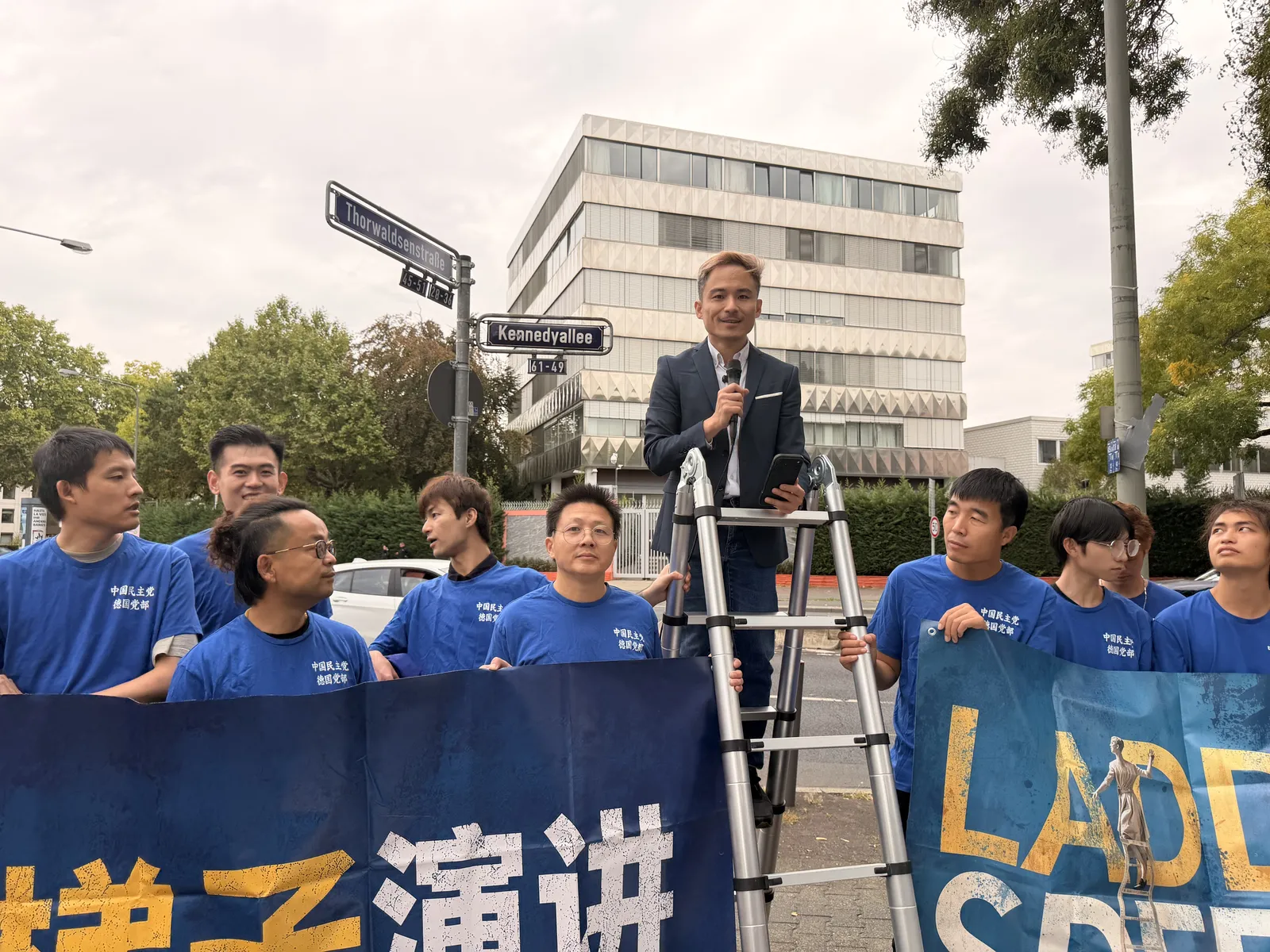 A speaker in a dark jacket addresses the consulate from the ladder while members in blue T-shirts stand below.
