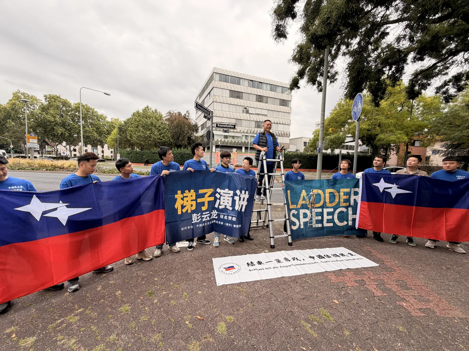 Members hold party flags on both sides of the banner, with a white banner on the ground in front of them and a speaker on the ladder.