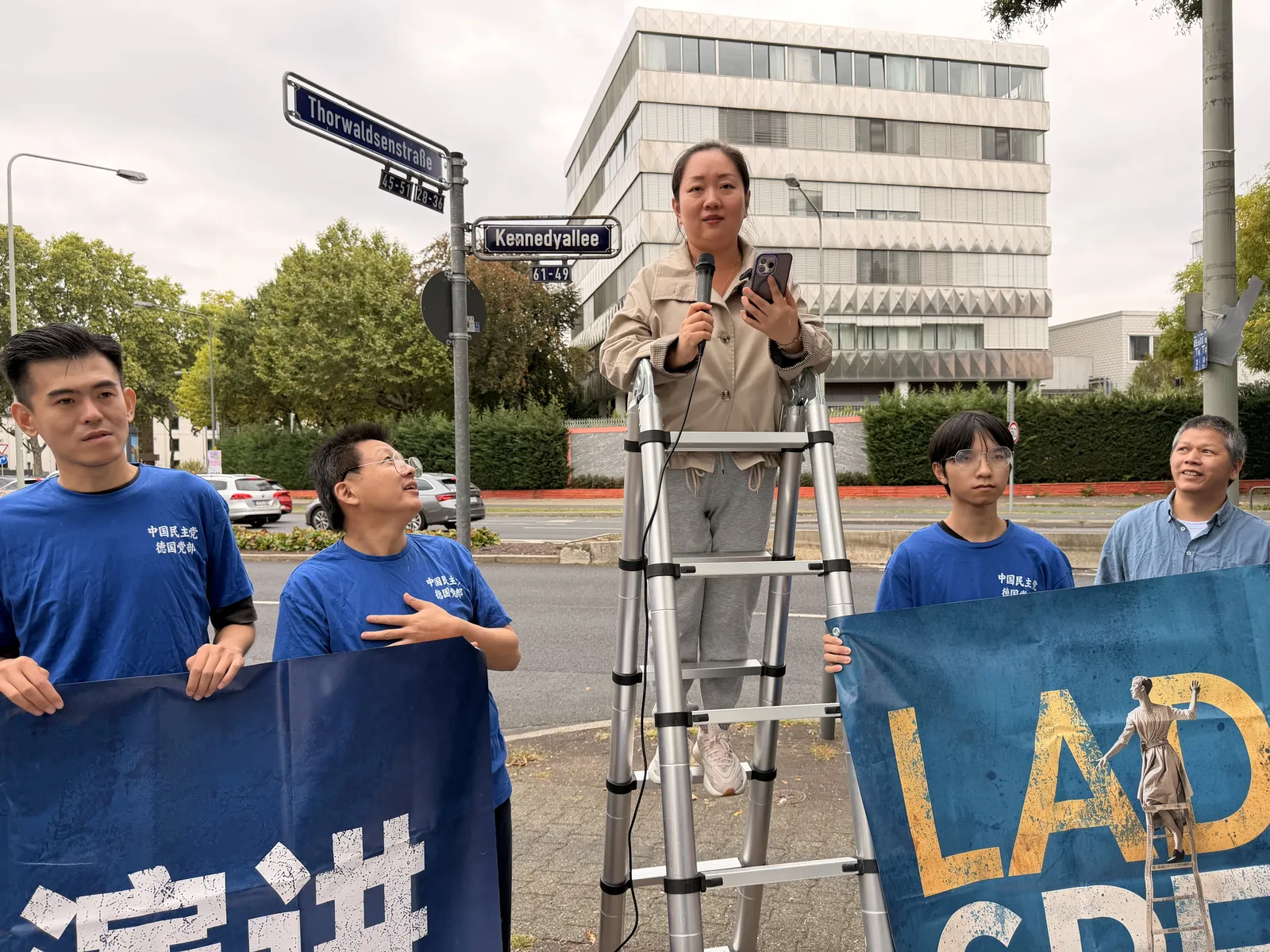 A woman speaks into a microphone on the ladder while members hold the banner.