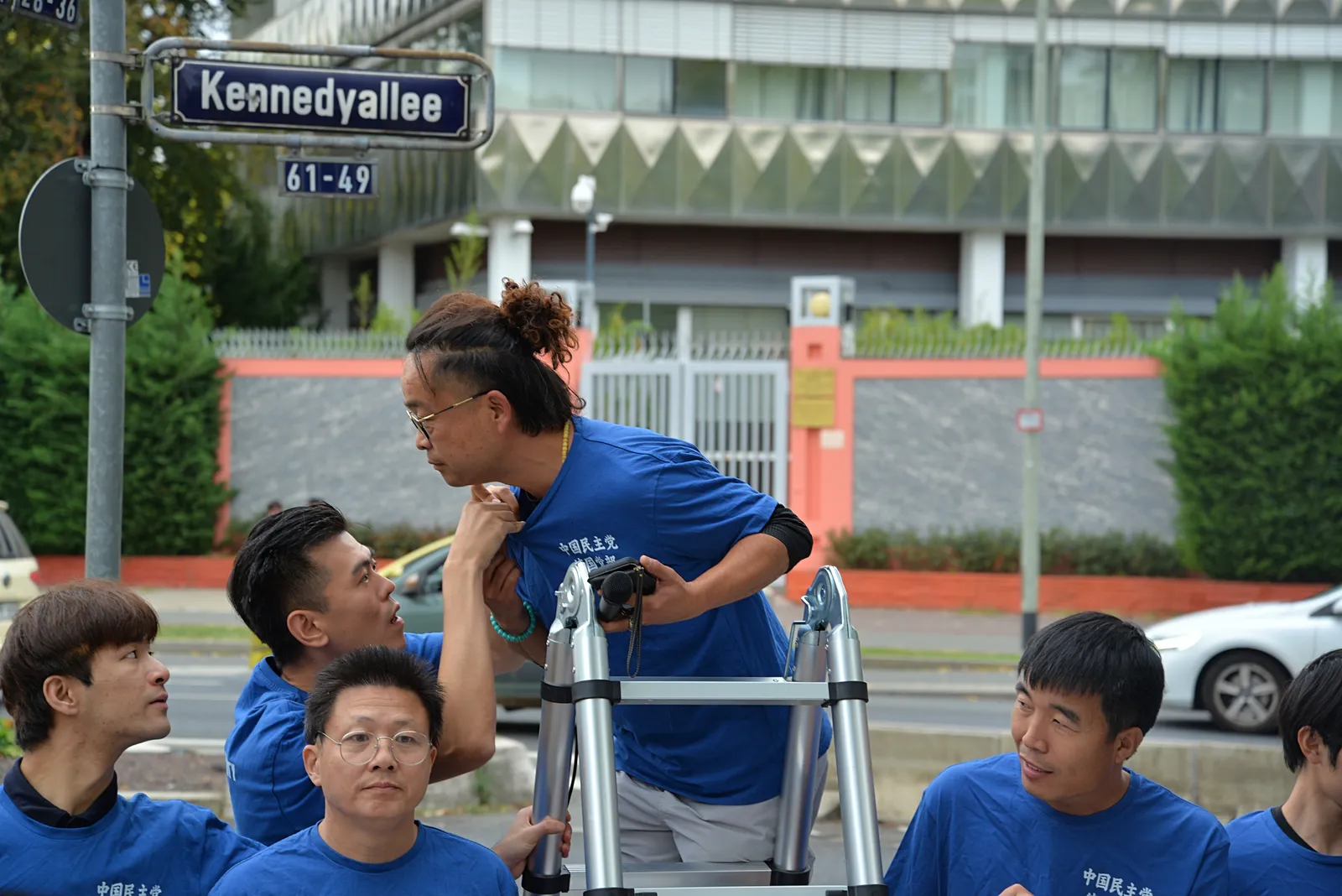 Members in blue T-shirts gather close around the ladder, with the Kennedyallee street sign and the consulate fence behind them.