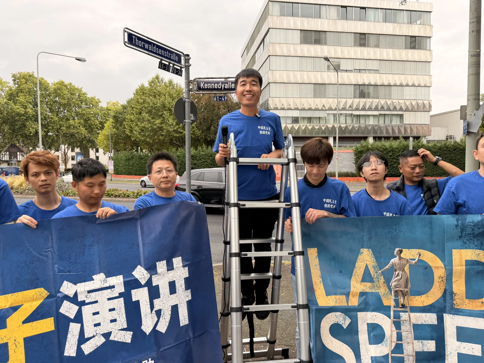 A speaker holds a microphone on the ladder, with the “LADDER SPEECH” banner and members below.