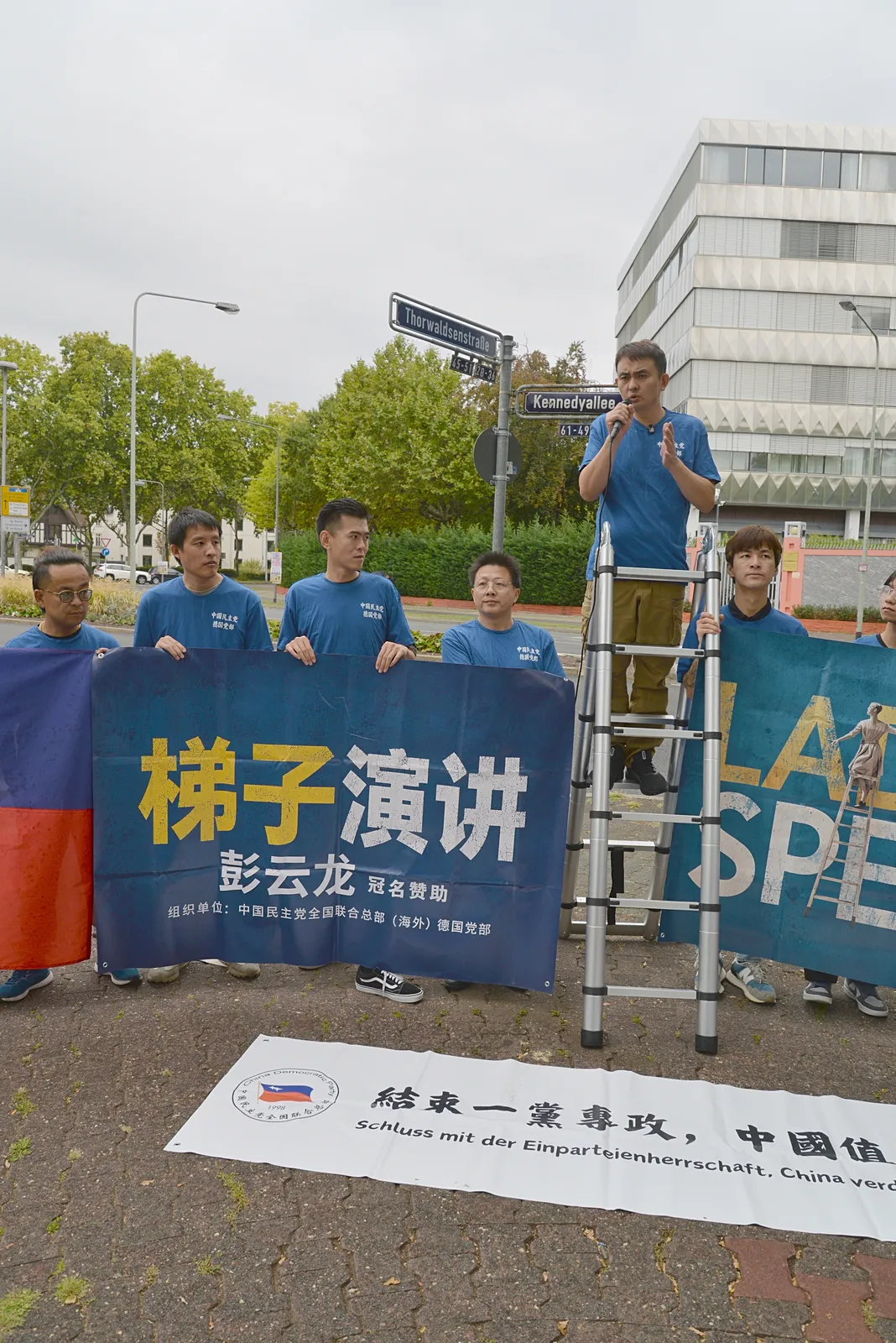 A wide view of members with flags and banners on the traffic island and a speaker on the ladder in the distance.
