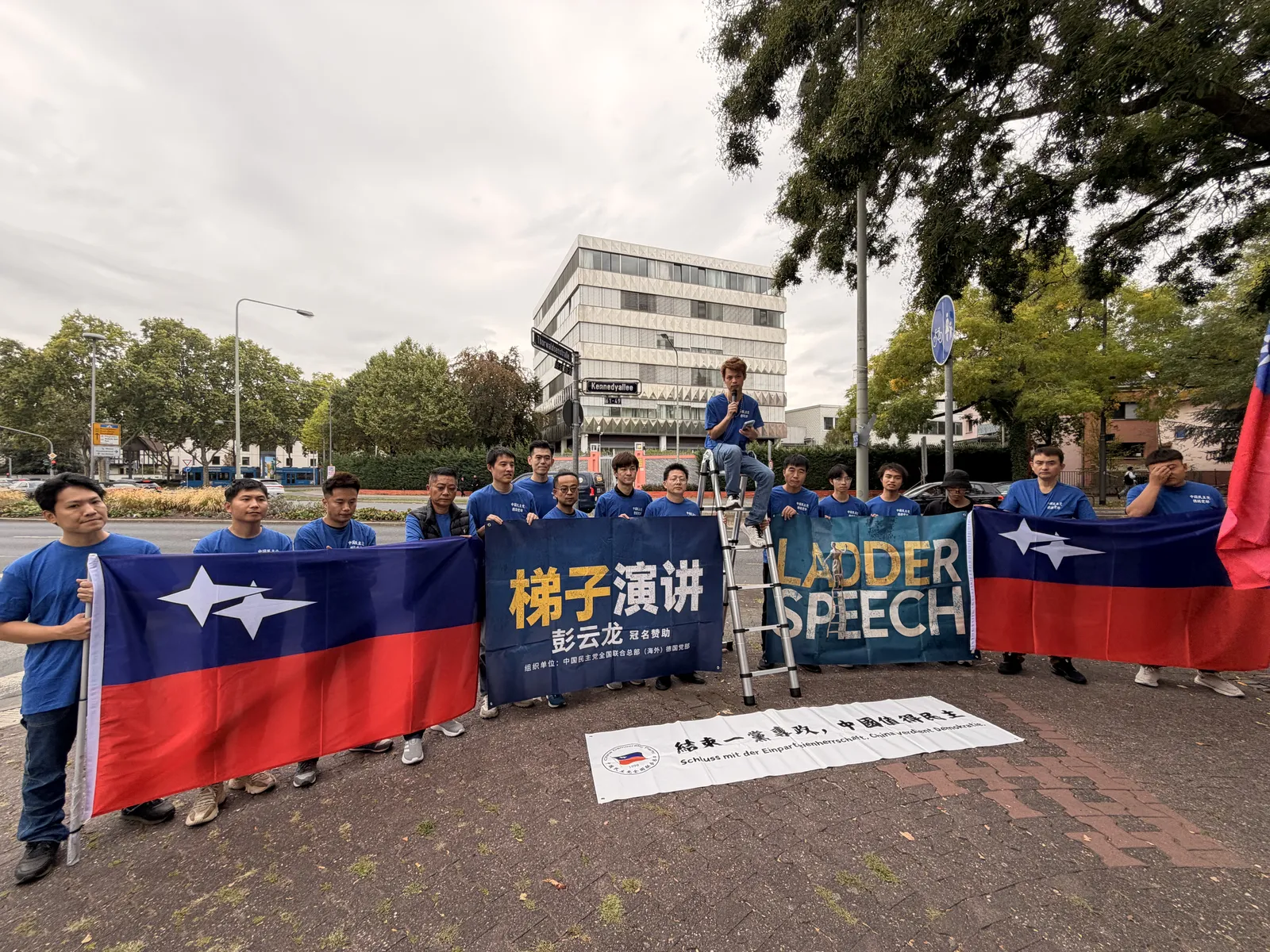 A wide view of the whole group with party flags, the banner and a white banner on the ground, across the street from the consulate.